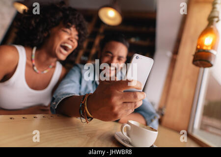 Young couple at cafe taking selfie using smart phone. Young man and woman laughing while taking self portrait in cafe. Stock Photo