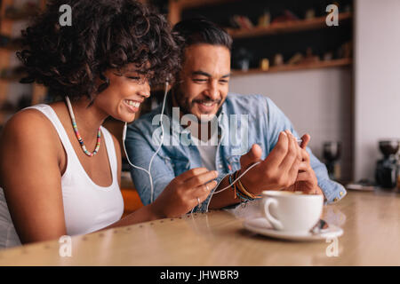 Happy young couple sitting at coffee shop having video chat on mobile phone. Young man and woman with earphones at cafe using smart phone. Stock Photo