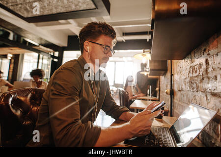 Handsome young man sitting at cafe with laptop and using smart phone. Casual businessman reading text message on his cell phone. Stock Photo