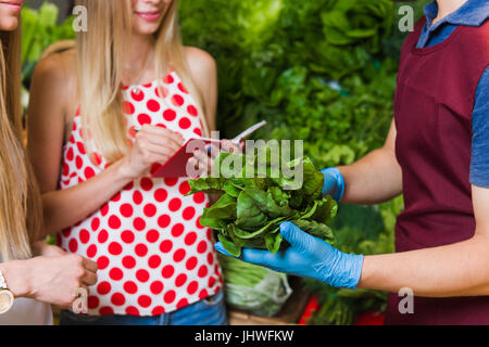 The seller and two blonde women at a vegetable market. Girlfriends buy vegetables on the market. Stock Photo