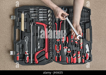 Black toolbox on the light tabletop in the workshop. Man with a tattoo holds a spanner above it. There are black-red pliers and screwdrivers, ratchets Stock Photo