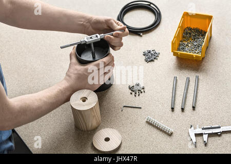 Man with tattoo is using a t-wrench on a black metal cylinder on the light table. On the table there are wooden billets, cable, plastic box with screw Stock Photo