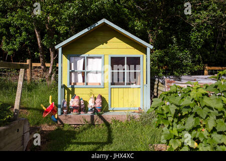 A small painted wood house for children to play in with gnome statues for decoration in a UK garden. Stock Photo