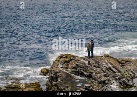 A young couple stand on wave-worn rocks looking out over the Atlantic Ocean at Bristol, Maine, USA. Stock Photo