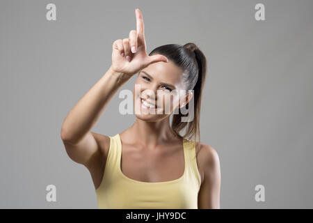 Young fit sporty woman showing letter L loser hand gesture on forehead smiling at camera over gray studio background. Stock Photo