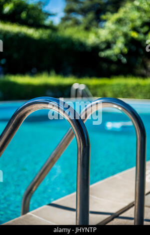 Swimming pool ladder close up, nobody in blue water and green garden out of focus in background Stock Photo