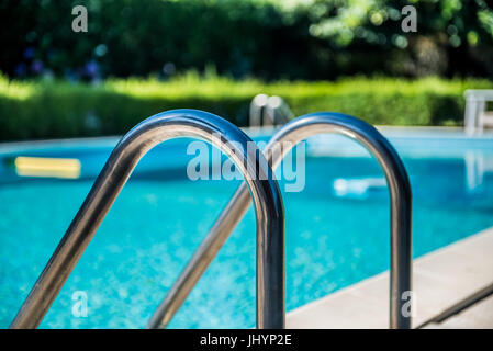 Swimming pool ladder close up, nobody in blue water and green garden out of focus in background Stock Photo