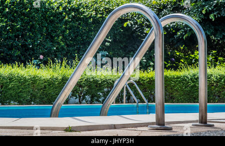 Swimming pool ladder close up, nobody in blue water and green garden out of focus in background Stock Photo