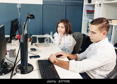 Couple of young designers working at modern office, two coworkers discussing fun project over a laptop, little team of businesspeople smiling and look Stock Photo