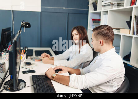 Couple of young designers working at modern office, two coworkers discussing fun project over a laptop, little team of businesspeople smiling and look Stock Photo