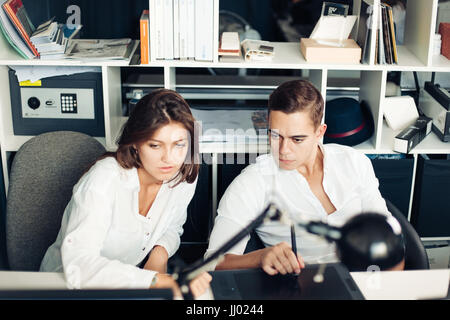 Couple of young designers working at modern office, two coworkers discussing fun project over a laptop, little team of businesspeople smiling and look Stock Photo