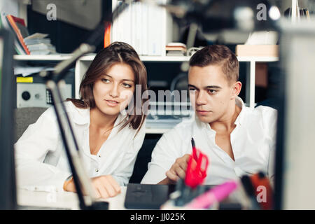 Couple of young designers working at modern office, two coworkers discussing fun project over a laptop, little team of businesspeople smiling and look Stock Photo