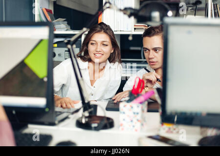 Couple of young designers working at modern office, two coworkers discussing fun project over a laptop, little team of businesspeople smiling and look Stock Photo