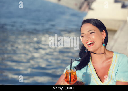 Beautiful smiling woman sitting at the seaside cafe,  drinking cocktail  and looking aside. Her hair blown by the wind. Copy space on the left side Stock Photo