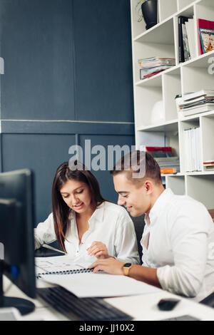Couple of young designers working at modern office, two coworkers discussing fun project over a laptop, little team of businesspeople smiling and look Stock Photo