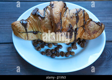 freshly baked big croissant with a chocolate filling and covered with chocolate on a white plate with coffee beans. low angle shot Stock Photo