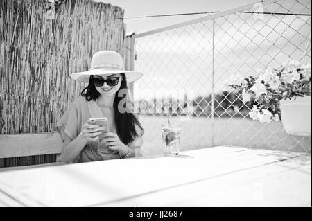 Sweet young lady wearing hat and sunglasses with a cocktail using her smartphone while sitting by the lake. Black and white photo. Stock Photo
