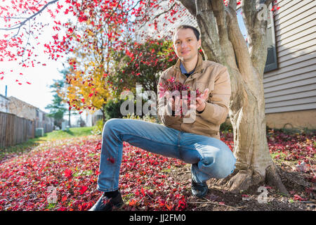 Young man sitting by tree in autumn red maple leaves foliage holding them in neighborhood smiling Stock Photo