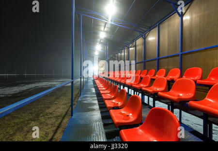 Empty tribune with rows wet red plastic seats on a football field with snow and puddles. (at night) Stock Photo