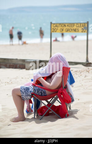 Man sitting on a beach with his head under a towel in hot weather, Boscombe, Bournemouth, Dorset, UK, 18th July 2017.