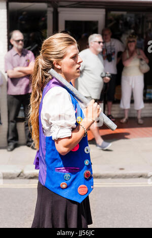 Young blonde Caucasian woman, 29s, Royal Liberty Morris Dancer standing in bright sunshine in the street, holding metal pole against shoulder. Stock Photo