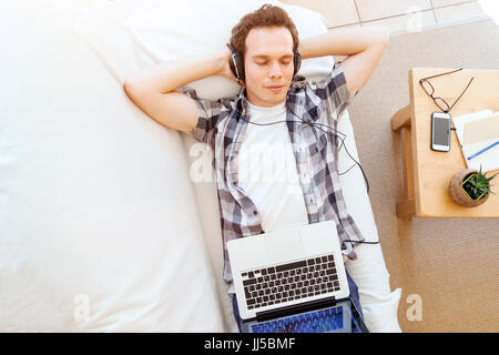 top view of young man in headphones lying down on the couch at home and enjoying listening music on computer Stock Photo