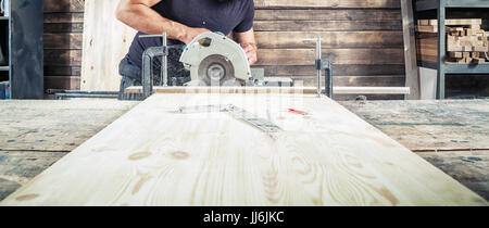 A young brunette man in a black T-shirt is sawing a tree with a modern circular saw in the workshop Stock Photo