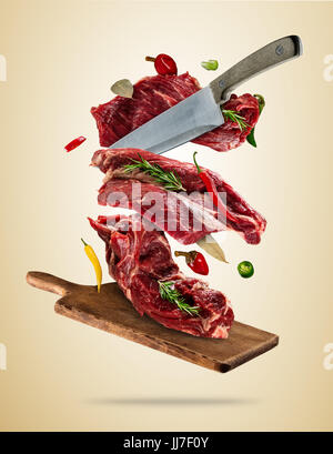Flying pieces of raw steaks, with ingredients for cooking, served on woodenboard. Knife cuting the meat. Concept of food preparation in low gravity mo Stock Photo