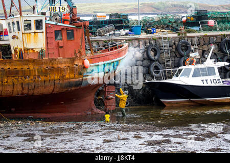 Dock scene, man pressure washing, a rusty, old fishing trawler, in Tobermory Harbour, Isle of Mull, Scotland. Stock Photo