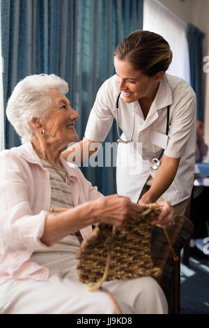 Smiling senior woman knitting while looking at female doctor in retirement nursing home Stock Photo