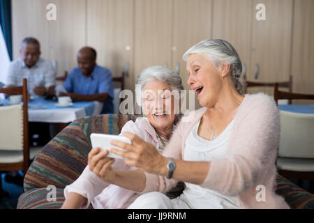 Senior friends taking selfie through smart phone while sitting on sofa at nursing home Stock Photo