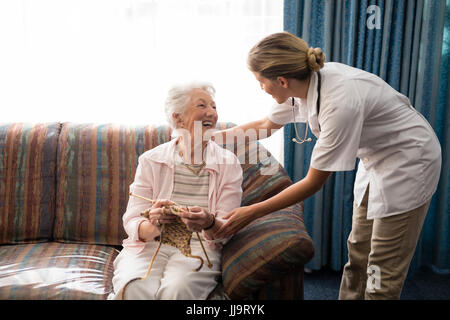 Cheerful senior woman holding knitting wool while looking at female doctor against window in nursing home Stock Photo