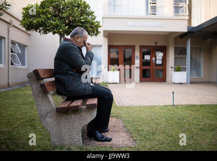 Side view of depressed senior man sitting on bench against retirement nursing home Stock Photo