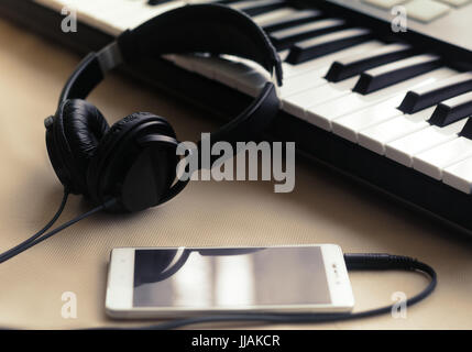 The headphones are connected to the phone lying next to the synthesizer on a beige background. Side view Stock Photo