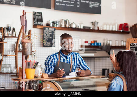Smiling young African barista talking to a customer and writing down an order in a notebook while standing at the counter of a cafe Stock Photo