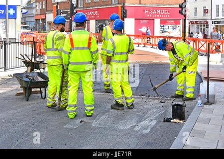 Group of workmen resurfacing town centre road one man using pick axe four others taking short break all wearing hard hats & full high visibility kit Stock Photo
