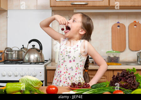 child girl eat cherries, fruits and vegetables in home kitchen interior, healthy food concept Stock Photo