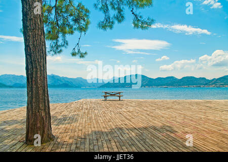 wooden table with benches on wooden plate platform at Aegean sea with big pine tree at foreground on sunny day in Marmaris, Turkey Stock Photo
