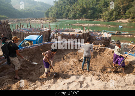 Loa men and women, working for SinoHydro, shovel sand on the shore of the Nam Ou River in Hat Sa, Laos. The sand is for making concrete used in the construction of Dam #6 being built upstream. Stock Photo