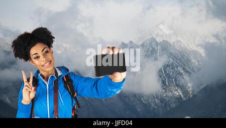 Digital composite of Hipster taking selfie and gesturing peace sign while standing against snowcapped mountains Stock Photo