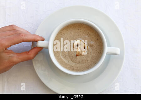 Woman hand holding white porcelain tureen pot of champignon mushroom cream soup in on the table, close up, high key, elevated top view Stock Photo