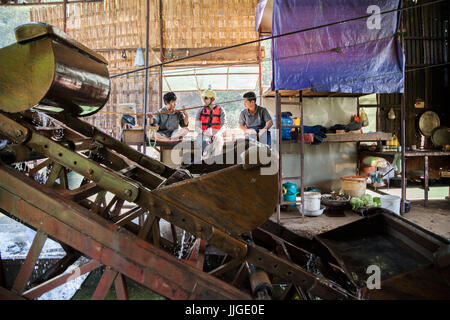Robert Hahn (center) chats with Vietnamese workers, while drinking a Vietnamese beer, on a mechanical gold dredge floating on the Nam Ou River, Laos. Six men live on the vessel for months, rotating throughout the day and working in pairs to keep the machine in constant operation. Stock Photo