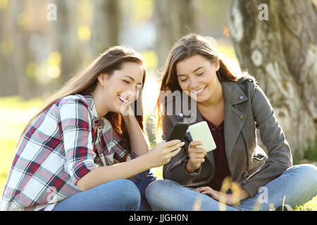 Two happy friends watching media content in their smart phones sitting on the grass in a park Stock Photo