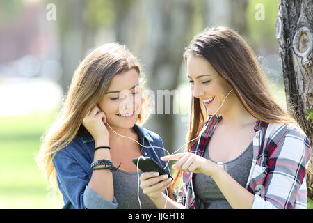 Two happy teen friends listening music sharing earphones and smartphone in a park Stock Photo