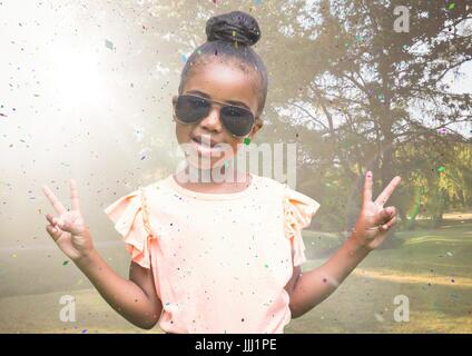 Girl in sunglasses making peace signs against blurry park with flare and confetti Stock Photo