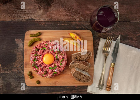 A photo of a steak tartare with a raw egg yolk, gherkins, capers, rye bread, roasted potatoes, a glass of red wine, a fork, and a knife, shot from abo Stock Photo