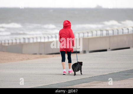 A woman dog walker walking along the coastal promenade at Rhyl in North Wales wearing a hooded jacket to  protect from wind and rain with her dog on a lead, Wales, UK Stock Photo