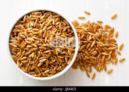 Kamut wheat kernels  in white ceramic bowl isolated on painted white wood from above. Spilled wheat. Stock Photo