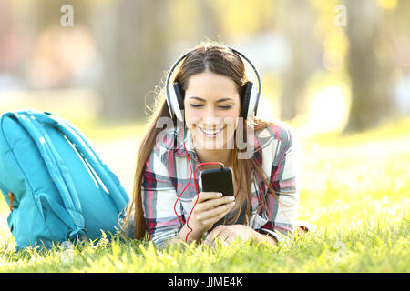 Happy student listening audio lessons on line with headphones and a smart phone lying on the grass in a campus Stock Photo