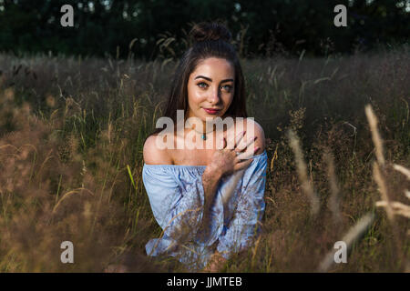 A pretty young woman is lit up in golden light whilst posing for a portrait in a meadow of wild grass near Liverpool before sunset. Stock Photo